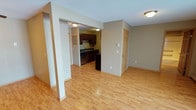 This single-floor space features a thoughtfully designed open layout that promotes a natural flow between the living and kitchen areas. Light hardwood flooring extends throughout the main living space, lending warmth and continuity to the interior. Neutral beige walls complement the light tones of the flooring, creating a bright and welcoming ambiance.

The kitchen is defined by rich, dark wood cabinetry that contrasts elegantly with the lighter surroundings. It is equipped with a black appliance suite and a sink positioned along the back wall, offering a practical and organized workspace. The kitchen is partially separated from the living area by a structural column and an arched passageway, adding architectural character while maintaining an open feel.

A paneled interior door leads to an additional room, providing functional separation within the unit. The textured ceiling adds subtle dimension overhead, while flush-mount lighting ensures consistent illumination throughout. The overall design balances practicality with a clean, understated aesthetic, making the space functional and comfortable for everyday living.