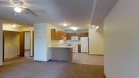 This single-floor residence offers an open-concept layout that seamlessly connects the kitchen and living areas, creating a cohesive and functional living space. The kitchen is equipped with warm honey-toned wood cabinetry that provides generous storage, complemented by white appliances including a refrigerator, electric range, and built-in microwave. A peninsula-style breakfast bar extends from the cabinetry, serving as both a food preparation surface and an informal dining or seating area. The kitchen floor features vinyl tile, transitioning naturally into the carpeted living area, which adds warmth and comfort underfoot. Ceiling-mounted flush lighting illuminates both areas evenly. The neutral color palette throughout — soft whites and warm beiges on the walls — contributes to a clean, understated ambiance. A partial wall separates the kitchen from the adjoining spaces, maintaining an open feel while subtly defining functional zones. A hallway visible in the background suggests additional rooms beyond the main living area. Overall, the space presents a practical and comfortable layout well-suited for everyday living.