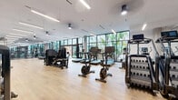 This fitness center in San Diego, CA offers a generously sized, single-floor exercise space designed to accommodate a comprehensive range of workout needs. The open-plan layout is finished with light hardwood flooring throughout, lending warmth to the contemporary aesthetic, while a crisp white ceiling fitted with linear LED lighting ensures bright, even illumination across the entire floor.

Floor-to-ceiling windows and large glass panel walls line the perimeter, flooding the interior with natural light and providing views of lush tropical greenery and an adjacent outdoor turf area, creating an energizing atmosphere. Full-length mirrors are strategically positioned along multiple walls, enhancing the sense of space and supporting proper form during exercise.

The cardio zone is well-appointed with treadmills, stair climbers, stationary bikes, and rowing machines, while the strength training area features a robust free-weight section with a full rack of dumbbells and barbells, power racks, cable machines, and a variety of selectorized strength machines. A dedicated functional training area includes TRX suspension systems, battle ropes, medicine balls, and balance equipment. Ceiling fans contribute to comfortable airflow throughout. The space also connects to an outdoor fitness area, extending workout possibilities beyond the interior.