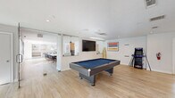 This thoughtfully designed single-floor space offers a versatile blend of recreational, collaborative, and productive environments across a generously sized footprint. The layout is anchored by a bright, open-plan main area featuring light wood-style flooring, recessed lighting, and an abundance of natural light from large windows throughout.

A central recreation zone is equipped with a full-size pool table, wall-mounted television, and an arcade game, creating an inviting social hub. Glass partition walls elegantly divide this area from a spacious conference room, which accommodates a long communal table surrounded by upholstered chairs — ideal for group meetings or collaborative work sessions.

Private offices and smaller meeting rooms offer carpeted flooring, recessed lighting, and natural window views of the surrounding landscape, providing quiet, focused work environments. A cozy lounge nook features a plush armchair, warm pendant lighting, and coastal-inspired décor, adding a relaxed touch to the space.

The kitchen area is finished with subway tile backsplash, light wood cabinetry, open floating shelves, stone-look tile flooring, and a large white countertop island — functional and visually appealing. Throughout the space, curated artwork and coastal accents, including decorative oars and vibrant framed prints, lend a cohesive, contemporary character to the overall design.