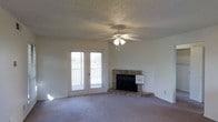 This single-floor space centers around a well-appointed living area designed for comfort and functionality. The room's most notable feature is a classic brick corner fireplace, adding warmth and a traditional aesthetic focal point to the space. Opposite the fireplace, a set of French doors floods the room with natural light while providing direct access to an outdoor balcony, offering views of the surrounding area. A ceiling fan with an integrated light fixture is centrally mounted to the textured popcorn ceiling, ensuring year-round air circulation and ambient lighting. The floor is finished with wall-to-wall carpet in a neutral gray tone, contributing to a cozy and cohesive atmosphere throughout. The walls are painted in a clean, light neutral shade, creating a bright and open feel that complements the natural light entering through the French doors. The overall layout is open and versatile, providing ample space for a variety of furniture arrangements. The combination of the brick fireplace, soft carpeting, and natural light creates a comfortable and inviting living environment.