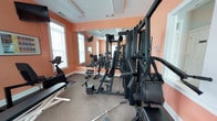 Located at 3952 Horseshoe Rd N in Little River, South Carolina, this single-floor fitness facility is designed to support a comprehensive workout experience. The gym room features warm peach-toned walls that create an energetic atmosphere, complemented by large windows that allow abundant natural light to fill the space. A full-length wall mirror spans one side of the room, visually expanding the area and allowing users to monitor their form during exercise. The facility is outfitted with a multi-station cable and strength training machine, providing a wide range of resistance exercises. A dedicated free weights section includes a dumbbell rack with an assortment of weights and an adjustable workout bench, catering to various strength training routines. A stationary exercise bike offers cardiovascular training options. The carpeted flooring provides a comfortable surface underfoot, while the well-maintained interior reflects a clean and functional environment. With its combination of natural light, mirrored walls, and diverse equipment, this fitness space delivers a practical and motivating setting for residents or members looking to maintain an active lifestyle.