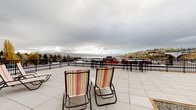 This expansive rooftop terrace offers a well-appointed outdoor retreat with sweeping panoramic views of a nearby waterway, a prominent bridge, and a city skyline visible in the distance. The terrace is finished with large-format light grey tiles that extend across the entire surface, creating a clean, cohesive aesthetic underfoot.

Multiple seating zones are thoughtfully arranged throughout the space. Striped fabric lounge chairs and reclining sun loungers are positioned to take full advantage of the waterfront views, while clusters of teak-slatted armchairs with dark metal frames provide additional casual seating areas suited for conversation. Dining tables with coordinating chairs are also present, accommodating outdoor meals or gatherings.

A decorative landscaping section adds greenery to the rooftop, featuring large charcoal-toned cubic planters filled with ornamental trees and gravel, set within a defined bordered bed. This greenery softens the hardscape and contributes visual interest to the overall design.

The perimeter is enclosed by a black metal railing that maintains open sightlines while ensuring safety. The terrace also includes a utility structure in one corner. Surrounding tree canopies and neighborhood rooftops are visible beyond the railing, further enriching the elevated outdoor setting.