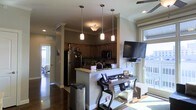 This single-floor residence offers a thoughtfully designed open-concept layout that seamlessly connects the kitchen and living areas, creating a cohesive and functional living environment. The kitchen is a standout feature, highlighted by warm wood cabinetry, granite countertops, and a peninsula with a built-in sink that serves as both a workspace and a casual gathering spot. Stainless steel appliances, including a full-size refrigerator and over-the-range microwave, add a modern touch. Three pendant lights hang gracefully above the peninsula, casting a warm, inviting glow over the space. Crown molding along the ceilings and a decorative column accent lend architectural character throughout. Rich, dark hardwood floors run continuously through the main living areas, grounding the space with warmth and elegance. Natural light pours in through large windows, offering views of an adjacent outdoor deck or balcony. A wall-mounted flat-screen television anchors the living area, which is complemented by a ceiling fan for year-round comfort. The overall ambiance is warm, modern, and practical — ideal for everyday living and entertaining alike.