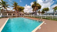 Located at 119 S Redland Rd in Florida City, Florida, this outdoor amenity area showcases a generously sized rectangular swimming pool as its centerpiece. The crystal-clear, turquoise water is framed by tile coping and a broad, paved sun deck that provides ample space for lounging and recreation. Pool depth markers are visible along the perimeter, indicating a gradual depth suitable for a range of swimmers. Lounge chairs positioned along the deck offer a comfortable setting for relaxation under the South Florida sun. A white picket fence encloses the entire pool area, providing both security and a clean, classic aesthetic. Lush tropical landscaping lines the fencing, with mature palm trees and dense greenery creating a verdant, resort-like atmosphere. An accessibility lift is present at the pool's edge, ensuring inclusive access for all users. Adjacent to the pool, a low-profile building with a dark shingled roof and covered entrance serves as a support facility, complemented by pathway lighting for evening use. The surrounding neighborhood, visible beyond the fence, features single-story residential buildings with matching rooflines. The overall setting evokes a relaxed, tropical ambiance characteristic of South Florida living.