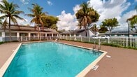 Located at 119 S Redland Rd in Florida City, Florida, this outdoor amenity area showcases a generously sized rectangular swimming pool as its centerpiece. The crystal-clear, turquoise water is framed by tile coping and a broad, paved sun deck that provides ample space for lounging and recreation. Pool depth markers are visible along the perimeter, indicating a gradual depth suitable for a range of swimmers. Lounge chairs positioned along the deck offer a comfortable setting for relaxation under the South Florida sun. A white picket fence encloses the entire pool area, providing both security and a clean, classic aesthetic. Lush tropical landscaping lines the fencing, with mature palm trees and dense greenery creating a verdant, resort-like atmosphere. An accessibility lift is present at the pool's edge, ensuring inclusive access for all users. Adjacent to the pool, a low-profile building with a dark shingled roof and covered entrance serves as a support facility, complemented by pathway lighting for evening use. The surrounding neighborhood, visible beyond the fence, features single-story residential buildings with matching rooflines. The overall setting evokes a relaxed, tropical ambiance characteristic of South Florida living.