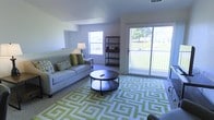 This single-floor living space offers a well-organized and inviting layout centered around a comfortable living area. The room is anchored by a light gray sofa dressed with green and patterned accent pillows, paired with a round dark-wood coffee table resting atop a bold green geometric-patterned area rug. The rug adds a striking visual element that ties the room's color palette together.

Natural light is a defining feature of the space, provided by a standard window on one wall and a large sliding glass door that opens to an outdoor area with visible greenery. The sliding door allows ample daylight to fill the room while offering a pleasant view of the surrounding landscape.

A dark metal open-shelf bookcase stands against one wall, offering both functional storage and a modern design accent. The neutral gray walls and standard ceiling height contribute to a clean, understated aesthetic throughout. The carpeted flooring adds warmth and complements the overall color scheme. The combination of natural light, outdoor access, and thoughtfully selected furnishings creates a functional and comfortable living environment.