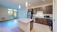Located at 350 Bishops Way in Brookfield, Wisconsin, this single-floor residence offers a thoughtfully designed layout encompassing one bedroom, one bathroom, a laundry area, a utility room, and two closets. The open-concept kitchen and living area serves as the heart of the home, featuring rich dark-stained cabinetry, granite countertops, and a full suite of stainless steel appliances including a range, microwave, dishwasher, and refrigerator. A central kitchen island with a white base and granite top provides additional workspace and casual dining potential, while pendant lighting and recessed fixtures illuminate the space. Light hardwood-style flooring runs throughout the main living and kitchen areas, lending warmth and continuity. The bedroom is a comfortable, well-proportioned retreat finished with plush carpeting, a ceiling fan, and a window that frames views of surrounding greenery, allowing natural light to fill the room. Two closets provide ample storage throughout the unit. The layout also includes a dedicated laundry area and utility room for added convenience. Neutral wall tones and clean finishes throughout the space create a bright, move-in-ready environment well suited for everyday living.