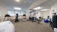 Located at 9300 Collegeview Rd in Bloomington, Minnesota, this fitness center offers a dedicated exercise room equipped to support a well-rounded workout routine. The space features patterned carpeted flooring throughout and is illuminated by fluorescent ceiling lights, creating a bright and functional environment. Large windows along one wall allow views into an adjacent interior atrium, adding an open and airy feel to the room.

The exercise room is outfitted with a solid selection of cardio equipment, including multiple treadmills, an elliptical trainer, and a stationary bike. Strength training needs are met with a multi-function cable machine, an adjustable weight bench, and a dumbbell rack. Stability balls round out the equipment offerings for flexibility and core work. A wall-mounted flat-screen television provides entertainment during workouts, while a water dispenser and informational bulletin board near the entrance add practical convenience.

Full-length mirrors are strategically placed to expand the sense of space and allow users to monitor their form. The textured ceiling and neutral wall tones contribute to a clean, no-frills atmosphere focused on functionality. This well-appointed fitness center provides residents with a convenient and comprehensive option for maintaining an active lifestyle without leaving the building.