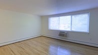 This single-floor space is designed with a clean and functional aesthetic, offering a welcoming atmosphere through its simple yet practical layout. The main living area is highlighted by light-toned hardwood floors that run throughout, adding warmth and a classic character to the space. Large multi-pane windows dominate one wall, allowing abundant natural light to fill the room while providing views of the surrounding greenery outside. The windows are fitted with horizontal blinds, offering adjustable light control and privacy. A wall-mounted air conditioning unit sits beneath the windows, ensuring climate comfort during warmer months, while a baseboard heating system runs along the perimeter of the room, providing efficient warmth in cooler seasons. The walls are finished in a neutral off-white tone, creating a bright and airy ambiance that complements the natural light. The smooth white ceiling contributes to the open feel of the room. Overall, the space presents a straightforward and practical environment, well-suited for personalization, with its neutral palette and durable flooring serving as a versatile foundation for various furnishing styles.