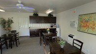 This single-floor space is designed for functionality and communal use, centered around a combined kitchen and dining area. The kitchen is outfitted with rich, dark espresso-toned upper and lower cabinetry that provides generous storage, complemented by light countertops and a tiled backsplash. A water dispenser and coffee maker are conveniently positioned on the counter, making the space well-equipped for everyday use. Overhead fluorescent lighting ensures the work area is well-lit, while a ceiling fan with integrated lighting helps maintain airflow throughout the room. The dining area accommodates multiple dark wood tables and matching chairs, arranged to support communal gatherings or group meals. Decorative potted plants placed atop the cabinetry and on the tables add a touch of greenery to the otherwise neutral, understated interior. The flooring is covered in a muted carpet, and the walls are finished in a clean, light tone. A wall-mounted heating and cooling unit provides climate control. The overall ambiance is straightforward and utilitarian, suited for a shared or multi-use environment such as a community room, breakroom, or similar common area.