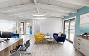 Located at 3252 SW 52nd Ave in Pembroke Park, Florida, this single-floor commercial office space offers a thoughtfully designed layout comprising three private offices, an open reception and lounge area, and a full bathroom. The standout architectural feature is the vaulted ceiling with exposed white-painted beams, which lends a sense of openness and airiness throughout the main area. Light wood-look tile flooring runs seamlessly across the space, complementing the neutral white and soft teal accent walls. The open reception area is bright and welcoming, furnished with a modern wood-tone reception desk, vibrant yellow and navy seating, and a cozy lounge arrangement that creates a professional yet inviting atmosphere. Large glass doors and clerestory windows allow natural light to filter in, enhancing the airy feel. One private office features a wall-mounted split AC unit and high clerestory windows, while another showcases a crisp white desk against a calming teal accent wall. The bathroom is finished with large-format wood-look floor tiles and matching wall tiles, recessed lighting, and clean, modern fixtures. This well-maintained office space is ideal for a small business seeking a polished and functional environment in South Florida.