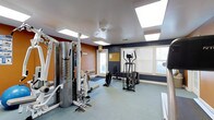 This single-floor fitness center offers a practical and comfortable environment for residents or members looking to maintain an active lifestyle. The space is designed with functionality in mind, featuring a combination of strength training and cardiovascular equipment, including a multi-station cable machine, an elliptical trainer, and additional cardio machines positioned near the windows.

The room is brightened by recessed skylight panels in the white ceiling, supplemented by natural light streaming through multiple large windows fitted with blinds along the navy blue accent wall. The contrast between the warm terracotta-toned wall and the deep navy blue creates a dynamic and energizing atmosphere well-suited for a fitness environment.

Durable, low-pile carpet flooring in a neutral grey-green tone covers the workout area, providing a stable and easy-to-maintain surface. A ceiling fan adds ventilation to keep the space comfortable during exercise. A bulletin board is mounted on the wall near the entry door, likely used for community notices or fitness guidelines. The layout provides adequate spacing between equipment, allowing for safe and comfortable use of each station. Overall, the fitness center is a well-organized, light-filled space designed to meet everyday exercise needs.