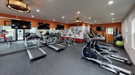 The fitness center at The Habitat @ Fort Collins is a thoughtfully designed, single-floor facility that combines functionality with an energetic aesthetic. The space is anchored by bold orange accent walls and a striking full-wall mural depicting colorful athletic silhouettes in dynamic poses, reinforcing the active atmosphere throughout.

The main workout area is generously proportioned, accommodating multiple treadmills, elliptical machines, and stationary cycling bikes arranged to allow comfortable spacing between users. A large wall mirror along one side visually expands the space and aids in form monitoring during exercise. Multiple wall-mounted flat-screen televisions are positioned throughout for entertainment during workouts.

A dedicated strength training zone features a cable machine, adjustable weight bench, medicine balls, stability balls, and a full dumbbell rack with an organized range of weights. Recessed lighting and a ceiling-mounted fixture provide consistent, well-distributed illumination across the space.

Natural light enters through windows on the perimeter walls, offering views of the surrounding outdoor greenery. A glass-paneled garage-style door serves as a standout architectural feature, flooding the space with additional daylight and providing a visual connection to the exterior. Durable rubber flooring throughout ensures safety and functionality. A restroom is accessible directly from the fitness area.