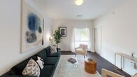 Located at 151 NE Second St in Oklahoma City, this single-floor residence features a well-considered layout with a cohesive modern design throughout. The interior is characterized by crisp white walls, creating a clean and airy backdrop across all rooms.

The living area is an inviting space with dark hardwood-style flooring, a large window dressed in sheer white curtains that allow natural light to filter through, and generous wall space suited for statement artwork. The room comfortably accommodates a full seating arrangement, lending itself to relaxed everyday living.

Adjacent to the living area, the kitchen is designed with a striking two-tone palette — white upper cabinetry contrasts sharply with dark lower cabinets and dark countertops. A peninsula with bar seating extends the counter space and provides an informal dining option. Pendant lighting above the peninsula adds a refined touch, while black appliances and a tile backsplash complete the functional yet stylish space.

The bedroom features carpeted flooring, a ceiling fan, and a wide window with outdoor views, filling the room with natural light. It provides ample space for a four-poster bed frame and supplemental furnishings.

The bathroom maintains the unit's consistent palette with dark countertops, black cabinetry, a large vanity mirror, and tile flooring.