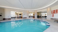 This indoor aquatic facility centers around a spacious, luminous swimming pool with crystal-clear blue water, accented by underwater lighting that creates a vibrant, inviting atmosphere. The pool is complemented by an adjoining hot tub, offering a complete aquatic experience. The space features a distinctive circular ceiling design with recessed lighting, contributing to the room's open, airy feel. Natural light streams in through large windows, brightening the neutral tones of the tile surround and light-colored walls. Stone-clad columns add a refined architectural detail, framing open views into adjacent lounge and dining areas furnished with tables and chairs. A mirrored wall on one side visually expands the space, while glass partitions maintain an open, connected feel throughout. The pool deck provides ample space for relaxed movement around the water. The overall ambiance is clean, modern, and welcoming, combining functional aquatic amenities with thoughtfully designed surrounding spaces suited for both leisure and socializing.