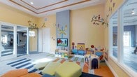 This single-floor children's play space is thoughtfully designed to spark imagination and encourage active play. The room is immediately eye-catching, with sunny yellow walls adorned with whimsical hand-painted murals depicting trains, birds, butterflies, and woodland scenes, creating an immersive, storybook-like environment. A central blue pillar serves as a focal point, decorated with a railroad crossing sign that adds a playful, thematic touch.

A wall-mounted flat-screen television is positioned above a low, white media console fitted with colorful, pull-out storage bins, providing both entertainment and organized toy storage within easy reach of children. The flooring features a vibrant, multi-colored patchwork area rug that defines the central play zone, complemented by durable hard flooring along the perimeter.

Soft, chartreuse upholstered ottoman cushions offer comfortable, low-to-the-ground seating for children. Along the walls, built-in play structures including a train-themed activity panel and interactive bead toys provide hands-on engagement. A mirrored doorframe near the entrance adds depth and light to the space. The tray ceiling with warm wood-toned molding trim lends a finished, polished quality to this lively and welcoming play environment.