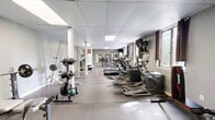 Located at 11 Farnsworth Dr in Slingerlands, NY, this dedicated fitness facility offers a comprehensive workout environment within a generously sized single-room layout. The space is designed to accommodate a wide variety of exercise modalities, making it suitable for both cardio and strength training routines.

The room features a durable low-pile flooring surface well-suited for gym use, neutral gray walls, and a dropped ceiling with recessed panel lighting that keeps the space consistently bright. Multiple windows along one wall, dressed with dark curtain panels, allow natural light to filter in while maintaining a focused workout atmosphere. A wall-mounted flat-screen television adds an entertainment option for cardio sessions.

Cardio equipment is prominently represented, with multiple Spirit treadmills and elliptical trainers positioned along the windowed wall. A stationary bike rounds out the cardio offerings. On the strength side, the space includes a rack of dumbbells reflected in a large wall mirror, barbell weight racks, a weight bench, and several cable and multi-function strength machines. The mirrored wall visually expands the room and allows users to monitor their form during exercise.

Structural support columns are integrated into the layout, and the floor plan flows openly from one equipment zone to the next, maximizing usable space throughout.