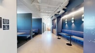 Located at 2506 W Colfax Ave in Denver, Colorado, this single-floor commercial suite offers a thoughtfully designed workspace that blends functionality with contemporary style. The layout includes two private offices and an open common area, all unified by a striking deep-blue and white color palette that creates a professional yet welcoming atmosphere.

The main corridor doubles as a lounge and reception zone, featuring built-in blue upholstered bench seating on both sides, accented by oval wall sconces and small black pedestal tables. The polished concrete flooring reflects light throughout, enhancing the sense of openness. Exposed white-painted ductwork and structural elements traverse the ceiling, lending an industrial character to the space.

One of the private offices serves as a conference room, equipped with a central worktable, ergonomic chairs, a wall-mounted display, whiteboard walls, and pendant lighting above a colorful geometric area rug. The other area transitions into a bright, sunlit lounge with large windows offering views of the Denver skyline, including a glimpse of a nearby bridge. Warm-toned seating, a concrete structural column, and abundant natural light give this zone a relaxed, collaborative feel.

A built-in blue cabinet nook provides integrated storage and workspace equipment. The overall design balances bold aesthetics with practical office functionality.