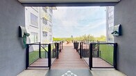 This thoughtfully designed outdoor amenity space is set within the interior courtyard of a multi-story residential building, offering a welcoming retreat above street level. The centerpiece is a generously sized composite wood deck, finished in a warm reddish-brown tone and accented by large, cylindrical planter pots that add greenery and structure to the layout. Adirondack chairs are arranged to take full advantage of the open-sky exposure and panoramic views of the surrounding urban landscape and distant skyline. String lights are strung overhead, creating an inviting ambiance for evening use. Flanking the deck are areas of lush artificial turf, providing a soft, low-maintenance contrast to the hardwood surfaces. The deck is accessed via a gated walkway equipped with modern black metal railings with wire mesh paneling, ensuring both safety and visual openness. Stairs lead down to ground-level courtyard access, while the surrounding multi-story facade — finished in a muted blue-gray palette with ornamental balconies and large windows — frames the space with architectural character. A utilitarian interior corridor with polished concrete floors and overhead fluorescent lighting provides functional access to the building's amenity level. Together, these elements create a versatile, well-appointed outdoor gathering space suited for both relaxation and socializing.