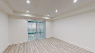 This single-floor unit presents a clean, contemporary design throughout. The main living area is generously proportioned, featuring light-toned wood-look flooring that extends seamlessly across the entire space, creating a cohesive and airy feel. Crisp white walls are complemented by recessed lighting, which provides consistent, even illumination. A tray ceiling detail adds architectural interest to the living area, while large sliding glass doors fitted with vertical blinds allow natural light to filter in and provide access to an outdoor balcony area.

The kitchen is well-appointed and efficiently laid out, featuring full-height dark wood-grain cabinetry with sleek bar-pull hardware, contrasted by light countertops and a stainless steel sink. A complete suite of stainless steel appliances includes a side-by-side refrigerator with water and ice dispenser, an electric range, an over-the-range microwave, and a dishwasher. Overhead ventilation is provided by a ceiling-mounted exhaust system.

Flanking the transition between the living and kitchen areas are tall, dark wood-grain storage cabinets that offer substantial built-in storage. The overall ambiance of the unit is modern, bright, and move-in ready.