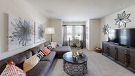 This single-floor residence offers a well-organized layout with distinct living, sleeping, cooking, and bathing areas, all finished in a cohesive neutral palette. The living room provides a comfortable, carpeted space centered around a sectional sofa, with a large window allowing natural light to fill the room and a textured ceiling adding subtle architectural detail. Wall art and an arc floor lamp contribute to the room's relaxed yet styled ambiance.

The kitchen follows a galley-style configuration, featuring dark espresso cabinetry paired with light granite-look countertops and hardwood-style flooring. Ample upper and lower cabinet storage runs along both sides, and the layout leads to an adjacent laundry or utility closet, adding functional convenience.

The bedroom is carpeted and bright, with multiple windows dressed in sheer curtains that allow diffused natural light. A ceiling fan with integrated lighting provides airflow and illumination, while the room comfortably accommodates a full bed and nightstand furnishings.

The bathroom is appointed with dark wood cabinetry, a single vanity with a granite-look countertop, a tall storage tower, and a tub-shower combination. Hardwood-style flooring, a chandelier-style light fixture, and framed wall art elevate the space with a polished, cohesive finish.