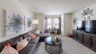 This single-floor residence offers a well-organized layout with distinct living, sleeping, cooking, and bathing areas, all finished in a cohesive neutral palette. The living room provides a comfortable, carpeted space centered around a sectional sofa, with a large window allowing natural light to fill the room and a textured ceiling adding subtle architectural detail. Wall art and an arc floor lamp contribute to the room's relaxed yet styled ambiance.

The kitchen follows a galley-style configuration, featuring dark espresso cabinetry paired with light granite-look countertops and hardwood-style flooring. Ample upper and lower cabinet storage runs along both sides, and the layout leads to an adjacent laundry or utility closet, adding functional convenience.

The bedroom is carpeted and bright, with multiple windows dressed in sheer curtains that allow diffused natural light. A ceiling fan with integrated lighting provides airflow and illumination, while the room comfortably accommodates a full bed and nightstand furnishings.

The bathroom is appointed with dark wood cabinetry, a single vanity with a granite-look countertop, a tall storage tower, and a tub-shower combination. Hardwood-style flooring, a chandelier-style light fixture, and framed wall art elevate the space with a polished, cohesive finish.