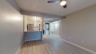 This single-floor residence offers a clean, functional layout with consistent light wood-tone vinyl flooring running throughout every room, providing a cohesive and low-maintenance living environment. The open living area serves as the heart of the home, featuring a ceiling fan, a large window fitted with horizontal blinds that allows natural light to fill the space, and a front entry door. The layout flows directly into the kitchen and hallway beyond.

The kitchen is efficiently designed with white upper cabinetry, gray lower cabinets, white subway tile backsplash, and bright white countertops. Stainless steel appliances — including a refrigerator, electric range, and over-the-range microwave — provide a modern finish. An electrical panel is integrated into the kitchen wall, and a peninsula counter separates the kitchen from the living area.

Adjacent to the kitchen, an in-unit laundry area accommodates a stacked or side-by-side washer and dryer setup. A hallway connects the main living space to the private areas of the home, which include two bedrooms. Each bedroom is finished in neutral warm-gray tones with vinyl flooring and windows with blinds offering natural light. The full bathroom features a single-sink vanity with a framed mirror, a toilet, and a shower-tub combination with a clean white surround.