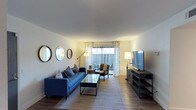 This single-floor residence offers a thoughtfully designed living space that balances comfort and modern style. The living room serves as the centerpiece of the home, featuring smooth, light-toned ceilings that contribute to an open and airy feel. Wood-style flooring runs throughout, adding warmth and continuity to the space.

The living area is well-lit, benefiting from natural light that streams in through large sliding glass doors fitted with sheer, patterned curtains. These doors provide direct access to an outdoor area, enhancing the sense of openness and connection to the exterior. A large flat-screen television is mounted on a sleek media console along one wall, while a bold blue tufted sofa and glass coffee table anchor the seating arrangement. Decorative round mirrors are arranged on the wall, adding visual interest and depth to the room.

A floor lamp and table lamp provide warm ambient lighting throughout the space. Neutral wall tones complement the furnishings and contribute to a calm, inviting atmosphere. The overall design aesthetic is clean and contemporary, with carefully chosen décor that creates a cohesive and livable environment suitable for both relaxing and entertaining.
