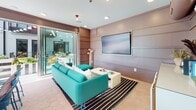 Located at 2904 W Clay St in Richmond, Virginia, this contemporary amenity space offers a well-designed blend of indoor comfort and outdoor living. The interior lounge area features a bold turquoise sofa, a wall-mounted flat-screen display, and large sliding glass doors that flood the space with natural light while providing seamless access to the outdoor courtyard. Warm-toned hexagonal tile flooring and horizontally paneled walls in muted mauve tones give the interior a modern, cohesive aesthetic. Adjacent to the lounge, a fully equipped kitchen showcases a generous white waterfall-edge island with vibrant blue bar stools, patterned cabinetry, full-size appliances, and a range hood — ideal for entertaining. Warm wood barn-style doors provide a stylish transition to additional rooms. The outdoor courtyard is a standout feature, enclosed by the multi-story building and framed by balconies with sleek metal railings. Lush raised planter beds filled with flowering plants and greenery surround a central seating area with a fire pit, complemented by cheerful yellow accent chairs. String lights overhead create an inviting atmosphere for evening gatherings. A putting green and additional al fresco dining areas complete this dynamic communal space.