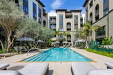 Located at 27665 Forbes Rd in Laguna Niguel, California, this beautifully designed outdoor amenity space offers a resort-inspired retreat within a contemporary multi-story residential complex. The centerpiece is a generously sized swimming pool with crystal-clear water, surrounded by wide stone-tiled pool decking and an array of cushioned chaise lounges and shaded umbrellas, perfect for relaxing in the Southern California sunshine. A separate spa pool, tucked into a lush, secluded corner accented by a natural stone wall and abundant tropical plantings, provides an intimate escape. The landscaping throughout is exceptionally lush, featuring towering palm trees, banana plants, olive trees, vibrant ground cover, and carefully curated tropical foliage that creates a verdant, resort-like atmosphere. Stylish outdoor seating areas with plush sofas, accent chairs, and decorative lanterns are arranged along tiled pathways between the greenery. Large floor-to-ceiling glass windows along the building's façade connect the interior clubhouse to the outdoor space, while arched entryways with ornate iron gates add an elegant architectural touch. The space is private, designated for resident use only, and reflects a high standard of design, comfort, and California outdoor living.