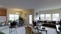Located at 300 Northwoods Rd in Middletown, NY, this single-floor residence offers a well-designed, open-concept layout that seamlessly connects the main living areas. The combined living and dining space is bright and airy, benefiting from an abundance of natural light streaming through multiple large windows that frame views of the surrounding neighborhood.

The living area is generously proportioned, accommodating a substantial sectional sofa and additional seating arranged around a glass-topped coffee table, creating an inviting space for relaxing or entertaining. The neutral, light-toned carpeting and soft wall colors contribute to a warm and welcoming ambiance throughout.

The dining area is defined by an elegant chandelier overhead and features a glass-top dining table with ornate metal chairs, adding a refined touch to the space. Lush potted plants bring natural elements indoors, enhancing the overall warmth of the room.

A glass-paneled entry door connects the main living area to the exterior, allowing additional light to filter through. The smooth, neutral-toned walls and consistent flooring throughout create a cohesive visual flow from one area to the next. Overall, this home presents a comfortable, light-filled environment with classic furnishings and a functional, open layout suited for everyday living.