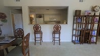 This single-story space showcases a practical and inviting open-concept layout, highlighted by a pass-through window connecting the kitchen to the living area. The kitchen is equipped with white cabinetry, a white refrigerator, and a built-in microwave above the range, all finished in a clean, neutral palette. The pass-through counter doubles as a breakfast bar, accompanied by two ornately carved wooden bar stools featuring decorative medallion backs and upholstered seats in a subtle patterned fabric, adding a touch of elegance to the functional space. The living area features neutral carpeting that complements the light-toned walls, creating a cohesive and understated aesthetic throughout. A carved wooden shelving unit on the right side of the room provides additional display and storage space. Partially visible on the left side of the frame is additional seating with matching carved wooden detailing, suggesting a coordinated furniture arrangement. The overall ambiance is calm and well-organized, with the open pass-through design enhancing the sense of connectivity between the kitchen and living spaces.