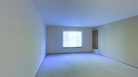 This single-floor residential space offers a clean and functional layout designed for comfortable everyday living. The main living area is generously proportioned, featuring wall-to-wall carpet flooring that adds warmth and softness underfoot. Neutral-toned walls provide a versatile backdrop, making the space easy to personalize and furnish to a variety of styles.

A large window serves as a focal point in the living area, allowing an abundance of natural light to fill the room and creating a bright, open atmosphere. The window also offers a view of the surrounding outdoor greenery, contributing to a pleasant and calm ambiance. Floor-level HVAC vents are positioned throughout, ensuring efficient climate control across the space.

The room connects to an interior door, suggesting access to additional areas of the unit such as a hallway, bedroom, or utility space. The ceiling height is standard and consistent throughout, reinforcing the open feel of the interior. Overall, the space presents itself as a well-maintained, move-in ready environment with a straightforward and practical design suited for a range of occupants.