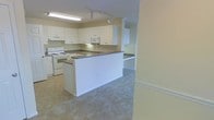 This single-floor space showcases an open-concept layout that seamlessly connects the kitchen and living areas. The kitchen is outfitted with white cabinetry that extends to the upper walls, providing generous storage. A built-in peninsula with a dark laminate countertop serves as a natural divider between the kitchen and the adjoining carpeted living area, offering additional prep space and a casual seating option. White appliances, including an electric range and microwave, complement the clean, bright aesthetic of the kitchen. Flooring transitions from vinyl tile in the kitchen to plush neutral carpet in the living space, defining each functional zone. Track lighting and a fluorescent ceiling fixture provide ample illumination throughout the kitchen. The walls are painted in a soft, warm neutral tone, contributing to the light and airy feel of the space. A built-in desk or nook is visible in the background, adding a practical workspace element. Overall, the space presents a functional, well-organized layout with a cohesive neutral palette suited for everyday living.
