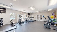 Located at 3900 Woodmere Park Blvd in Venice, Florida, this fitness center offers a comprehensive, single-floor workout facility designed to accommodate a wide range of exercise routines. The space is generously proportioned, providing ample room for both cardio and strength training activities without feeling overcrowded.

The cardio section is well-stocked with multiple treadmills, elliptical trainers, and stationary bikes, including Matrix-brand equipment. A dedicated strength training area features a multi-station cable machine, a weight bench, and a full dumbbell rack. An oversized wall mirror spans a significant portion of the room, enhancing the sense of space and allowing users to monitor their form during workouts.

The facility is brightly lit with recessed and flush-mounted ceiling fixtures, and natural light filters in through several windows and glass-paneled French doors that open to an adjacent outdoor area, hinting at a pool nearby. The durable rubber flooring provides traction and cushioning throughout. Wall-mounted televisions offer entertainment, while built-in shelving units near the entrance provide convenient storage. Ceiling fans assist with ventilation, contributing to a comfortable training environment. The clean, neutral-toned walls and well-maintained equipment give the space a professional and welcoming atmosphere.