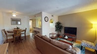 This single-floor residence offers a thoughtfully arranged open-concept layout that seamlessly connects the main living areas. The living room is comfortably furnished with a generous leather sectional sofa, a sleek flat-screen television on a wooden media console, and a decorative indoor tree that adds a natural touch. Hardwood-style flooring runs throughout, contributing to a warm and cohesive aesthetic, while a textured popcorn ceiling adds a classic residential character.

A partial dividing wall separates the living area from the kitchen zone, which features a raised breakfast bar with bar stools — ideal for casual dining or entertaining. The kitchen is equipped with standard appliances, including a refrigerator, and benefits from natural light streaming in through a nearby window. An adjacent dining area with a wooden table and chairs provides additional seating options.

The open flow between the living, dining, and kitchen spaces makes the layout feel expansive and functional. Neutral wall tones, including a subtle taupe accent wall in the living area, create a calm and inviting ambiance. Ceiling vents indicate a central HVAC system, ensuring year-round comfort. Overall, the space presents a practical and welcoming environment suited for everyday living.