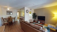 This single-floor residence offers a thoughtfully arranged open-concept layout that seamlessly connects the main living areas. The living room is comfortably furnished with a generous leather sectional sofa, a sleek flat-screen television on a wooden media console, and a decorative indoor tree that adds a natural touch. Hardwood-style flooring runs throughout, contributing to a warm and cohesive aesthetic, while a textured popcorn ceiling adds a classic residential character.

A partial dividing wall separates the living area from the kitchen zone, which features a raised breakfast bar with bar stools — ideal for casual dining or entertaining. The kitchen is equipped with standard appliances, including a refrigerator, and benefits from natural light streaming in through a nearby window. An adjacent dining area with a wooden table and chairs provides additional seating options.

The open flow between the living, dining, and kitchen spaces makes the layout feel expansive and functional. Neutral wall tones, including a subtle taupe accent wall in the living area, create a calm and inviting ambiance. Ceiling vents indicate a central HVAC system, ensuring year-round comfort. Overall, the space presents a practical and welcoming environment suited for everyday living.