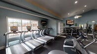This fitness center is organized into distinct workout zones, offering a comprehensive range of equipment for both cardiovascular and strength training. The cardio area houses a row of True treadmills positioned along a windowed wall, allowing natural light to fill the space. Wall-mounted flat-screen TVs are positioned for viewing during workouts, and a ceiling fan provides additional ventilation. An open shelving unit offers storage for personal items or equipment accessories.

The strength training area features vaulted ceilings illuminated by recessed lighting and ceiling fan fixtures, creating an airy and open atmosphere. Full-length mirrors line one wall, visually expanding the space while allowing users to monitor their form. This section is equipped with cable machines, a leg press, and various resistance machines. A dedicated free weights room provides a Smith machine, a weight bench, a barbell, and additional resistance equipment, with diamond-plate wall paneling and rubber flooring for safety and durability. A water fountain is conveniently accessible near the strength zone. Throughout, the space features cool blue-gray walls, dark rubber flooring, and a clean, functional design aesthetic.