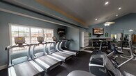 This fitness center is organized into distinct workout zones, offering a comprehensive range of equipment for both cardiovascular and strength training. The cardio area houses a row of True treadmills positioned along a windowed wall, allowing natural light to fill the space. Wall-mounted flat-screen TVs are positioned for viewing during workouts, and a ceiling fan provides additional ventilation. An open shelving unit offers storage for personal items or equipment accessories.

The strength training area features vaulted ceilings illuminated by recessed lighting and ceiling fan fixtures, creating an airy and open atmosphere. Full-length mirrors line one wall, visually expanding the space while allowing users to monitor their form. This section is equipped with cable machines, a leg press, and various resistance machines. A dedicated free weights room provides a Smith machine, a weight bench, a barbell, and additional resistance equipment, with diamond-plate wall paneling and rubber flooring for safety and durability. A water fountain is conveniently accessible near the strength zone. Throughout, the space features cool blue-gray walls, dark rubber flooring, and a clean, functional design aesthetic.