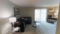This single-floor residential space offers a comfortable and functional layout centered around a well-appointed living area. The living room features plush wall-to-wall carpeting that adds warmth underfoot, complemented by neutral-toned walls that create a calm and inviting atmosphere. A large sliding glass door dressed with vertical blinds serves as the focal point of the room, allowing an abundance of natural light to pour in while providing access to the outdoors. The space is furnished with a dark leather sectional sofa adorned with decorative pillows, paired with a sleek glass-top coffee table that adds a contemporary touch. A stylish floor lamp and additional side table provide both ambient lighting and practical surface space. A bold piece of wall art adds a splash of character and visual interest to the room. The textured ceiling adds a subtle architectural detail throughout. Overall, the living area strikes a balance between comfort and style, offering a relaxed setting well-suited for both everyday living and entertaining.