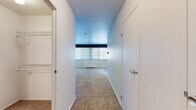 Located at 12540 Edgewater Drive in Lakewood, Ohio, this single-floor unit offers a practical and well-organized layout. The main living area is generously proportioned, featuring warm neutral carpeting that extends throughout the space, crisp white walls, and a large window dressed with vertical blinds that invites natural light and provides a bright, open atmosphere. A baseboard heating unit runs beneath the window, ensuring year-round comfort. The transition from the carpeted living space to light wood-look flooring marks the entry into the kitchen area, which is equipped with a black refrigerator and overhead cabinetry, offering functional storage and a clean aesthetic contrast. A hallway connects the living area to the remaining rooms, flanked by two closets that provide ample storage — one of which features a wire shelving system visible from the corridor. The bathroom is finished with white subway tile wainscoting, wood-look tile flooring, a white vanity with brushed nickel hardware, a mirrored medicine cabinet, and a full mirror above the sink, creating a clean and cohesive look. Throughout the unit, the consistent use of white tones and neutral finishes contributes to a simple, functional, and easy-to-maintain living environment.