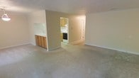 This single-floor space features an open-concept living area finished with wall-to-wall carpeting in a neutral tone, creating a clean and functional foundation. The living area is bright and spacious, with white walls and white trim that contribute to an airy, understated ambiance. A built-in pass-through counter with wooden cabinetry below connects the living area to the kitchen, providing a convenient serving or workspace while maintaining a sense of openness between the two areas. The kitchen is accessed through a dedicated doorway and is outfitted with wooden cabinetry, a white range, and a black oven, along with what appears to be tile or vinyl flooring. A hallway visible beyond the kitchen leads to additional areas of the unit. The overall design is practical and straightforward, with consistent neutral tones throughout. White baseboards frame each room, adding a subtle finishing detail. The layout is efficient, making good use of the available space on a single level.