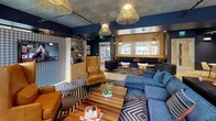 This single-floor amenity lounge is a thoughtfully designed social space that blends industrial and contemporary aesthetics throughout. The exposed concrete ceiling serves as a unifying architectural element, complemented by bold navy blue and warm gold accent walls that create a dynamic, energetic atmosphere. Striking woven pendant light fixtures hang overhead, casting warm light across the various seating zones below.

The main lounge area is anchored by a large navy sectional sofa and a pair of cognac leather wingback chairs arranged around a geometric-patterned area rug and a wood-topped coffee table. A houndstooth-patterned accent wall provides a bold backdrop for a wall-mounted flat-screen television, flanked by open wood shelving. A secondary seating zone features a navy built-in bench and modern accent chairs arranged around a chevron-patterned rug, with an additional flat-screen television mounted within a built-in media unit.

The galley-style kitchenette is a standout feature, showcasing a dramatic metallic mosaic tile backsplash, dark granite countertops, wood cabinetry, and under-counter beverage refrigerators. Large windows along one wall allow generous natural light to filter in, with views extending toward an outdoor pool area.