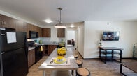 This single-floor unit offers a practical and comfortable layout designed to accommodate multiple occupants. The kitchen is a functional centerpiece, featuring rich dark wood cabinetry, light countertops, and a full suite of black appliances including a refrigerator, electric range, and over-the-range microwave. Wood-look flooring runs throughout the kitchen and common areas, adding warmth to the space. A stainless steel kitchen island with bar stools provides additional seating and serves as a casual dining area, complemented by a pendant light overhead.

The living room is a welcoming common space with wood-look flooring, a large window that brings in natural light, and ample room for multiple seating arrangements. The space connects naturally to the kitchen and dining area, creating an open, communal feel.

The unit includes multiple bedrooms, ranging from single-occupancy rooms with twin beds and built-in storage to larger rooms accommodating full-sized beds. Each bedroom features carpeted floors, a window with outdoor views, and dedicated study or side table space.

Two bathrooms serve the unit, each clean and well-appointed with a vanity, large mirror, and wood-look or tile flooring. The overall design is modern and straightforward, with neutral walls and consistent finishes throughout, making this a highly functional shared living space.