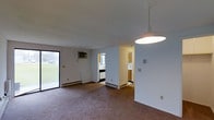 This single-floor residence offers a straightforward and functional layout designed for comfortable everyday living. The main living area is finished with wall-to-wall carpeting and features a large sliding glass door that invites natural light and provides direct views of the surrounding greenery outdoors. A wall-mounted air conditioning unit and baseboard heating ensure year-round climate control throughout the space. A pendant light fixture is centrally positioned, providing overhead illumination to the open living area. The kitchen is situated just off the main living space, separated by a partial wall that maintains an open feel while defining each zone. The kitchen is equipped with wooden cabinetry, a window that allows for additional natural light, and ample counter space for food preparation. Neutral white walls throughout the unit create a clean and versatile backdrop, making the space easy to personalize. The layout flows efficiently between the living and kitchen areas, offering a practical and cohesive living environment suitable for a range of occupants.