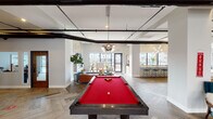 This single-floor amenity space is thoughtfully designed to offer a dynamic and inviting environment for socializing and recreation. The game room serves as a central highlight, featuring a bold red billiard table set atop striking herringbone-patterned flooring. Exposed black ductwork and industrial-style pendant lighting contrast elegantly with the bright, clean walls, lending the space a modern-industrial character. A stone-accented fireplace and comfortable lounge seating create a warm gathering area nearby. Large floor-to-ceiling windows throughout flood the space with natural light and offer views of the surrounding neighborhood. The lounge and dining area showcase an eclectic mix of colorful, sculptural pendant lights and a wicker hanging chair, adding a playful, artistic touch. A dedicated bar area with teal counter stools and pendant lighting provides an ideal setting for casual socializing. A coffee station with warm wood cabinetry and a professional espresso machine offers a welcoming spot for daily refreshment. The lobby area features a bold red structural column paired with lush indoor plants, patterned carpeting, and mid-century modern seating, creating a distinctive and energetic entryway. Throughout the space, curated artwork, varied textures, and thoughtful lighting combine to deliver a lively yet comfortable atmosphere.