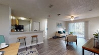 This single-floor residence offers a thoughtfully designed open-concept layout that seamlessly connects the kitchen, dining, and living areas. Wood-look vinyl plank flooring runs throughout the main living spaces, providing a clean and cohesive aesthetic, while textured ceilings and neutral gray walls create a calm, understated backdrop throughout.

The kitchen is equipped with warm-toned wooden cabinetry offering generous storage, a light-toned countertop with a double-basin sink, and black appliances including a range and dishwasher. A breakfast bar with room for barstools bridges the kitchen to the open living and dining area, making it ideal for casual dining or entertaining.

The living room is bright and inviting, featuring a ceiling fan and a large sliding glass door dressed with vertical blinds that allows abundant natural light and provides access to an outdoor area with greenery views.

The bedroom is a comfortable retreat with carpet flooring, a textured ceiling, and dual windows that frame outdoor views and fill the room with natural light. A walk-in closet with wire shelving offers practical storage. The bathroom features a full tub-shower combination with white tile surround, complemented by wood-look flooring for a consistent design throughout the home.
