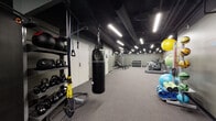 Located at 156 S Main St in downtown Akron, Ohio, this fitness facility offers a comprehensive single-floor workout space designed to accommodate a wide range of training styles. The interior features a dark, industrial aesthetic characterized by an exposed black ceiling with visible ductwork and structural elements, illuminated throughout by bright linear LED strip lighting that ensures consistent visibility across the entire floor.

The space is organized into distinct functional zones. A cardio section houses True-brand treadmills and an elliptical trainer, positioned along one wall for focused endurance training. Adjacent to this area, a strength training zone includes a power rack loaded with weight plates, a tiered dumbbell rack, and multiple adjustable benches suited for a variety of exercises. A dedicated functional training area features a Century heavy punching bag, TRX suspension straps, a battle rope, and kettlebells arranged on a freestanding rack. Stability and mobility training are also well-supported, with a collection of exercise balls, foam rollers, and yoga mats stored on a vertical rack.

The flooring throughout consists of durable carpet-style material suited for gym use, while the olive-toned walls and concrete block accents reinforce the utilitarian character of the space. A wall-mounted television and a water fountain add practical amenities to the facility.