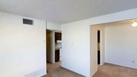 This single-floor space features an open and functional layout with a neutral color palette of soft white walls that create a clean, understated ambiance. The main living area is finished with wall-to-wall carpeting and includes a floor-level baseboard vent, contributing to the room's practical design. A textured popcorn ceiling runs throughout, adding a classic residential character to the space.

Visible from the main living area, the kitchen is accessible through an open doorway and features dark wood cabinetry along the upper and lower sections, paired with a white freestanding range and a range hood above. The kitchen's layout appears compact yet organized, making efficient use of its available space.

A second doorway on the opposite side of the main room leads to an additional area, likely a hallway or secondary room, also finished with the same dark cabinetry visible along one wall, suggesting possible storage or utility use. A wall-mounted thermostat and light switch plate are noted in the living area, reflecting standard residential conveniences. Overall, the space offers a straightforward and practical design well-suited for everyday living.